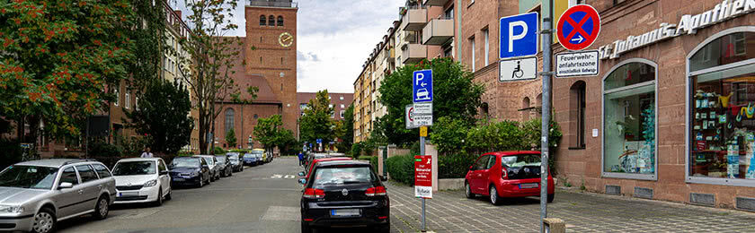 Wohngebiet mit mehreren parkenden Fahrzeugen. Ein rotes Fahrzeug parkt unerlaubt vor einer Apotheke auf dem Gehweg.
